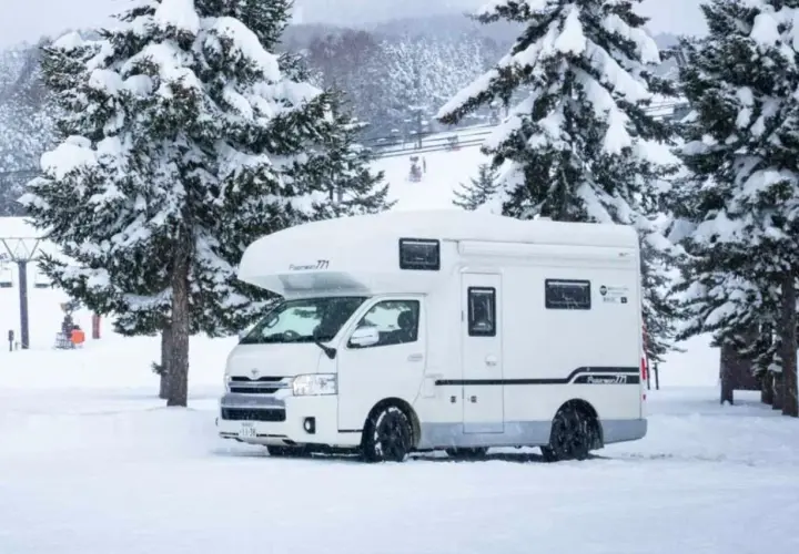 Camping car japonais blanc garé dans la neige, entouré de sapins enneigés. Des remontées mécaniques sont visibles en arrière-plan.
