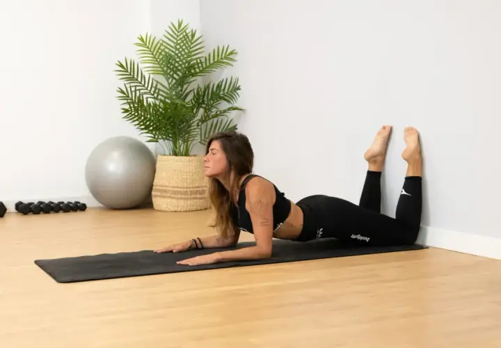 Mujer haciendo **pilates de pared gratis** en una esterilla, con pesas y pelota de ejercicio al fondo.