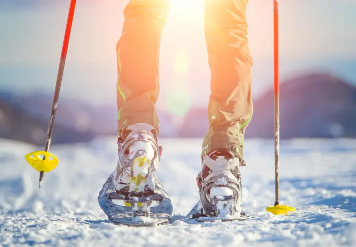 Piernas con raquetas de nieve y bastones listos para excursiones en la nieve bajo un sol brillante.