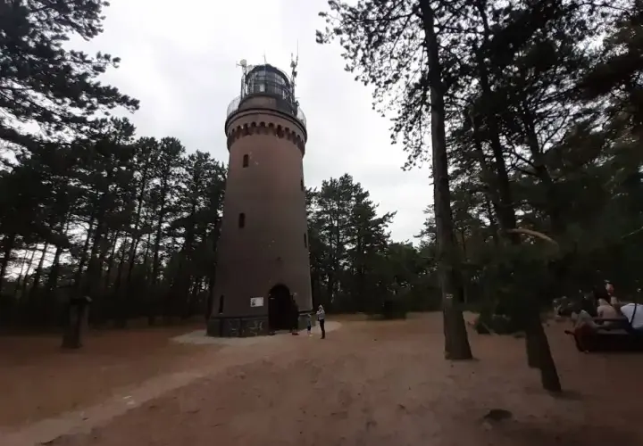 Latarnia morska w Czołpinie, otoczona sosnowym lasem, góruje nad piaszczystym terenem. W oddali widać fragmenty wydm.