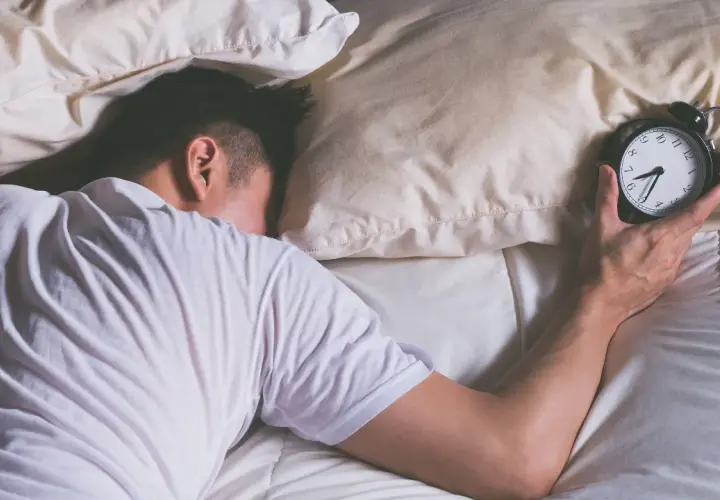 Mann schläft im Bett, Wecker liegt daneben. Wie viel Schlaf braucht man wirklich?