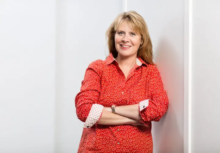 Petra Weber, eine lächelnde Frau mit blonden Haaren, trägt ein rotes Hemd mit Blumenmuster und hat die Arme verschränkt.