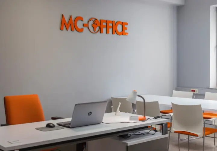 Pomarańczowe logo MC-OFFICE na szarej ścianie nad biurkiem z laptopem.