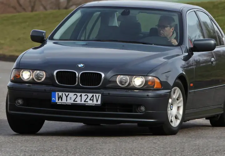 Ciemnoszary sedan BMW E39 z kierowcą w okularach jedzie po drodze.