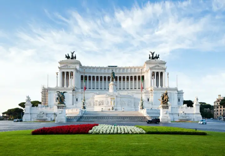 Il Vittoriano, monumento imponente, si erge maestoso. Vicino alla fermata metro Altare della Patria, un prato verde e fiori rossi e bianchi creano un contrasto vibrante.