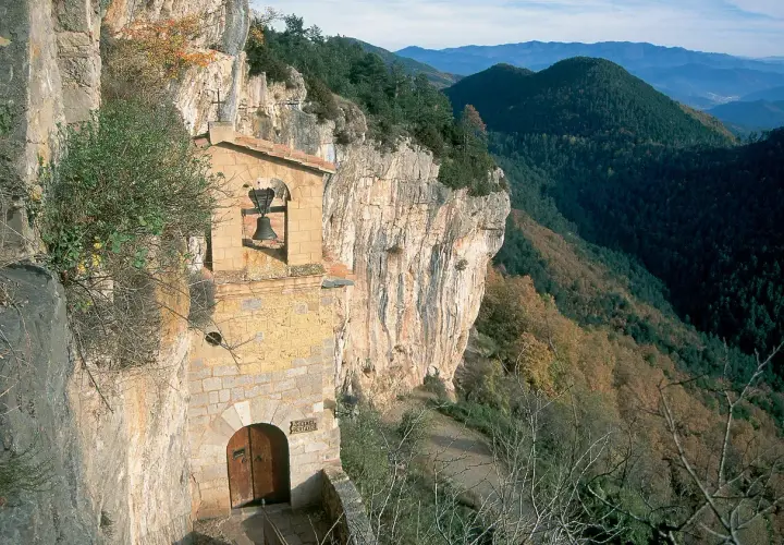 Ermita en la costa pubilla, aferrada a un acantilado rocoso con vistas a un valle boscoso. Una campana cuelga sobre la entrada.
