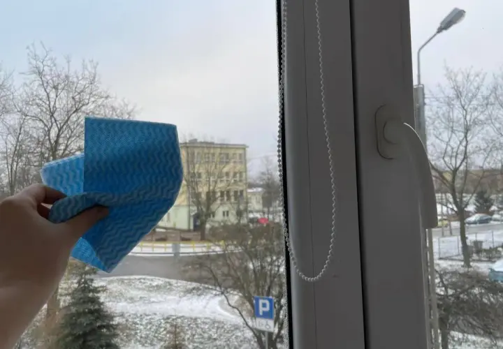 Ręka trzyma niebieską ściereczkę, gotową do mycia okna. Za szybą zimowy krajobraz z drzewami i budynkiem. Czy można myć okna na mrozie?