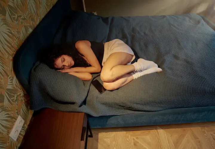 En ung kvinna sover oroligt på en säng, hennes andning tycks ansträngd, kanske av ångest. En mobiltelefon ligger bredvid henne.
