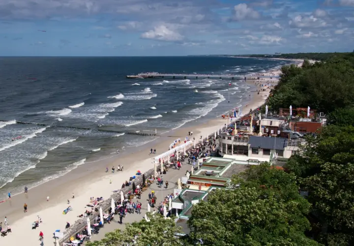 Tętniąca życiem promenada nad morzem, z plażą pełną ludzi, kawiarniami i molo w oddali.