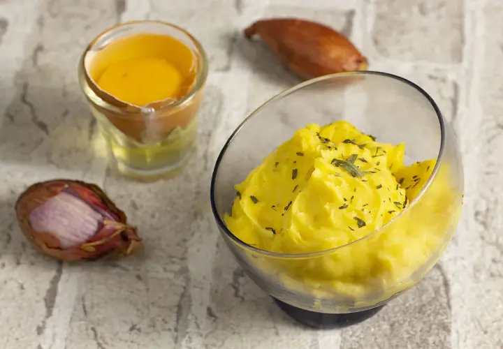 Una ciotola di salsa bernese cremosa, guarnita con erbe aromatiche, accanto a un tuorlo d'uovo e scalogni.