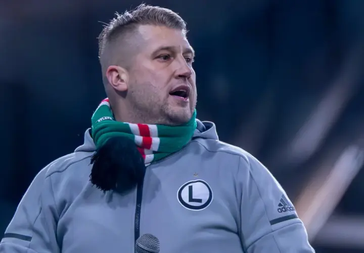 Staruch z Legii w szaliku klubowym, z mikrofonem przed ustami, gotowy do przemowy.