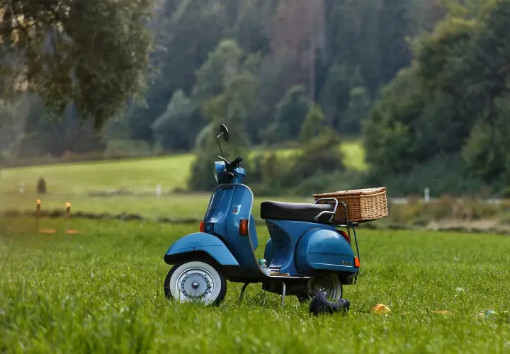 Niebieska Vespa z wiklinowym koszem na bagażniku, zaparkowana na trawie. Wygląda jakby czekała na przegląd skutera po terminie, gotowa na letnią przygodę.