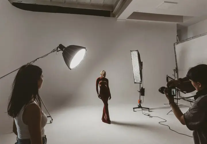 Modelka w czerwonej sukni pozuje na planie zdjęciowym. Fotograf z aparatem i asystentka przygotowują się do sesji.