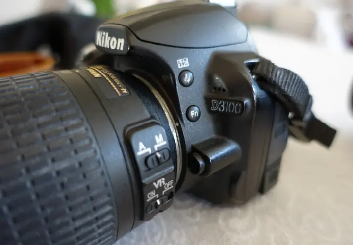 Zbliżenie na aparat Nikon D3100. Widoczny obiektyw z pierścieniem ostrości i przełącznikami AF/M oraz VR ON/OFF. D3100 to model, którego nikon d3100 dane techniczne warto poznać.