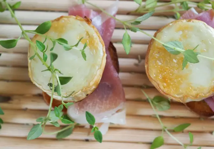 Dos bocaditos con crujiente de queso de cabra fundido y jamón serrano, adornados con hierbas frescas.