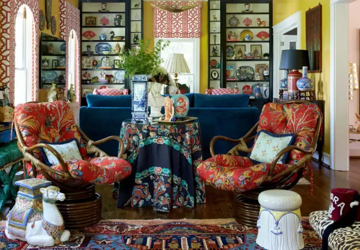 A maximalist living room bursts with color and pattern. Two rattan chairs with vibrant red floral upholstery face a navy sofa. Shelves overflow with decorative plates and vases.