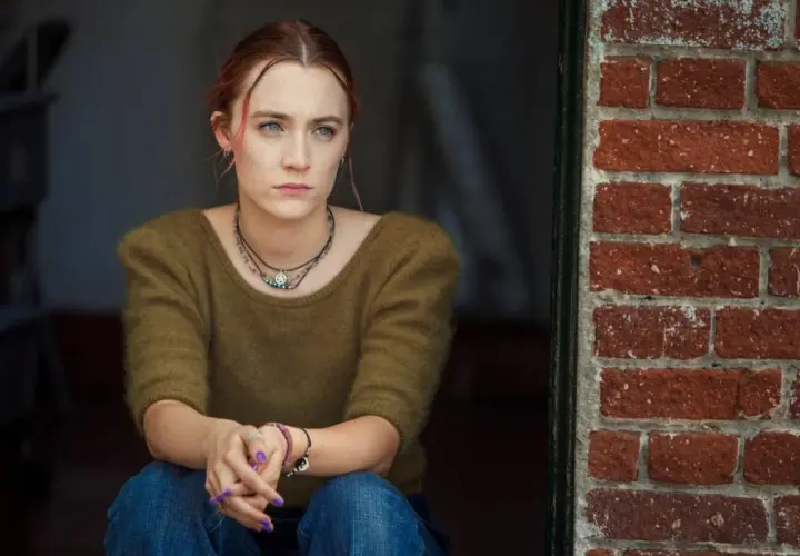Ragazza pensierosa, con maglione verde e jeans, seduta vicino a un muro di mattoni rossi. Una scena che ricorda la recensione di Lady Bird.