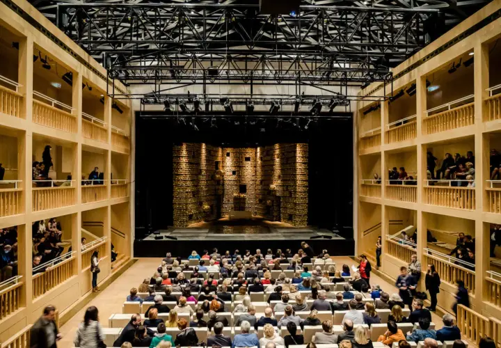 Widownia i scena w nowoczesnym teatrze królewskim w starej oranżerii. Drewniane galerie otaczają salę, a widownia zajmuje miejsca przed sceną.