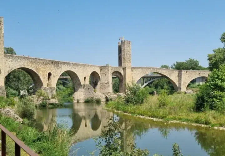 Puente medieval de Besalú, parte de una ruta histórica. Reflejo en el río y vegetación exuberante.