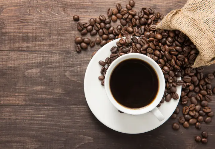 Aromatic coffee beans spill from a burlap sack onto a wooden table, next to a white cup filled with dark coffee. Poczuj kofeina działanie!
