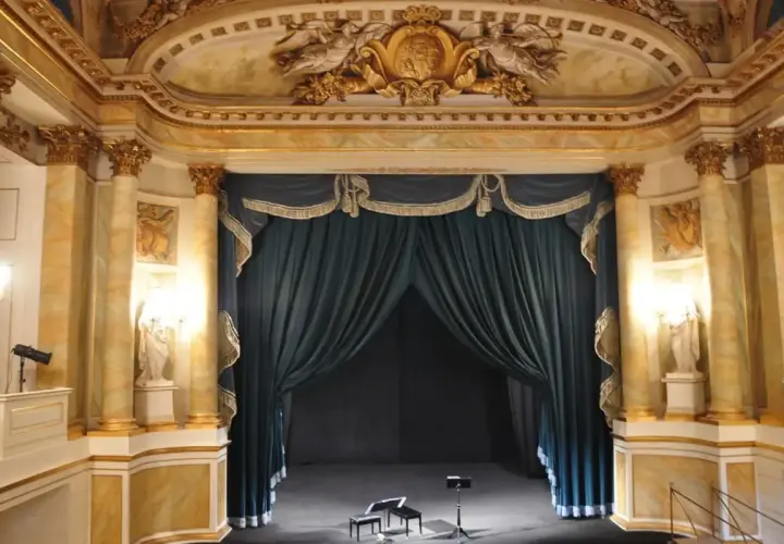 Złote zdobienia i bogate zasłony tworzą majestatyczne oświetlenie sceny teatralnej. Na scenie dwa stołki i pulpit nutowy czekają na artystów.