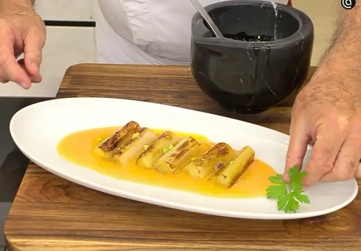 Puerros a la plancha, dorados y tiernos, servidos en una salsa anaranjada y decorados con perejil fresco.