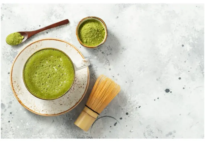 Jak pić herbatę matcha? Na stole leży spieniona zielona herbata w filiżance, obok bambusowa miotełka, łyżeczka z proszkiem i miseczka z matchą.