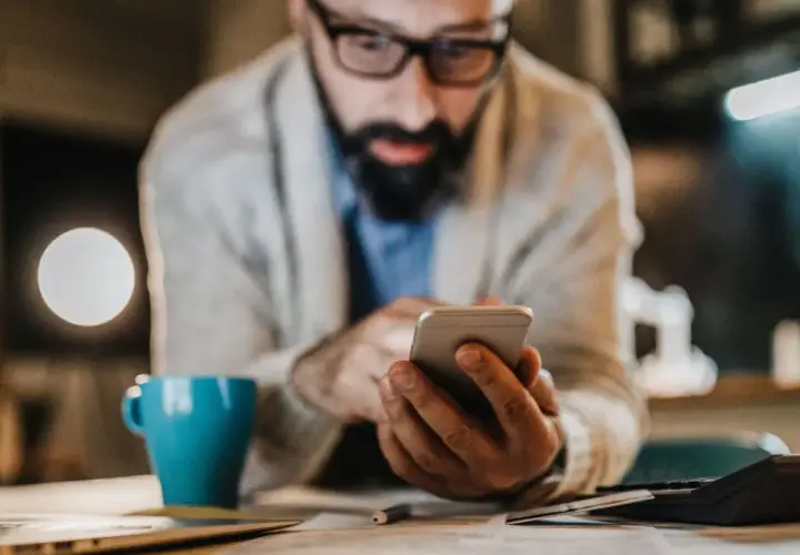 Mann mit Brille und Bart tippt auf Smartphone. Neben ihm eine blaue Tasse und ein Taschenrechner. Er sucht vielleicht, was ist die bankleitzahl.