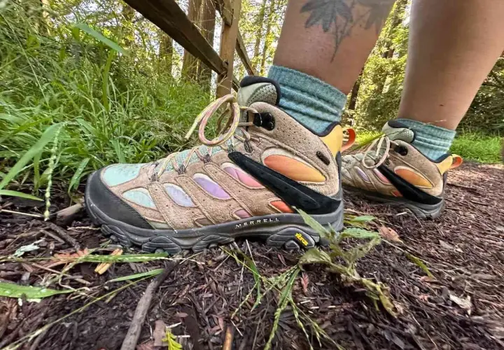 A person's feet wearing tan Merrell hiking boots with colorful accents and teal socks on a forest trail. These boots fit snugly, offering support for the hike.
