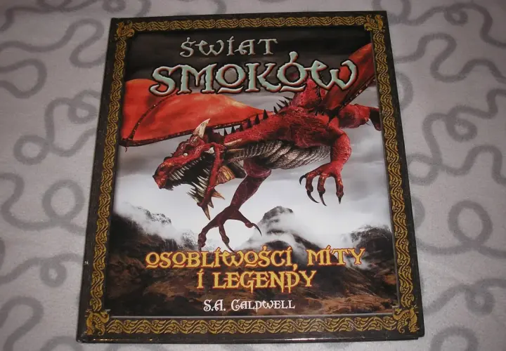 Okładka książki o smokach z czerwonym, groźnym stworzeniem na tle gór. Tytuł: "Świat Smoków: Osobliwości, Mity i Legendy".