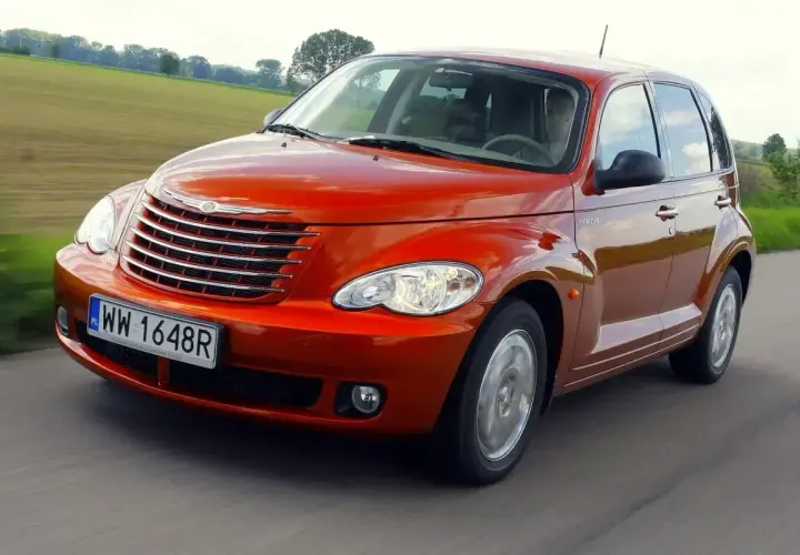 Pomarańczowy Chrysler PT Cruiser jedzie po drodze, za nim zielone pola i drzewa.