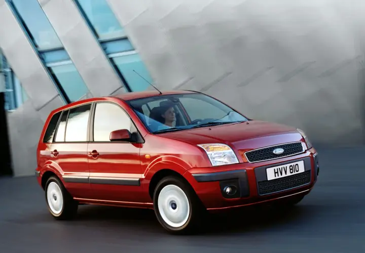 Czerwony Ford Fusion 1.4 benzyna w ruchu, z kobietą za kierownicą, na tle nowoczesnego budynku.
