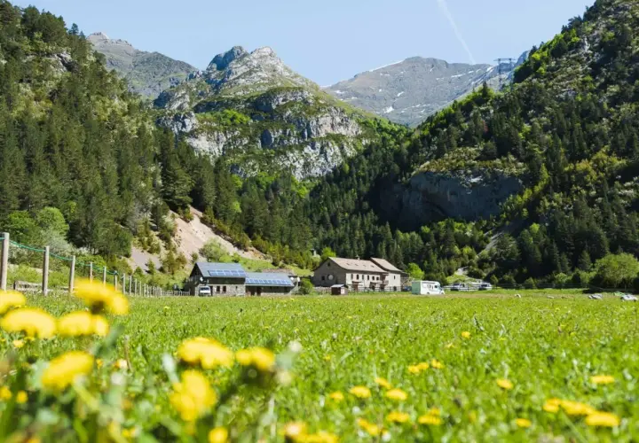 Prado florido con el Vignemale desde Bujaruelo al fondo. Casas rurales y montañas verdes bajo un cielo azul.