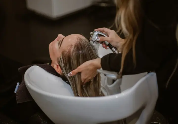 Una persona se lava el pelo con agua fría en un lavacabezas. El agua cae sobre su cabello rubio mientras una mano lo sujeta.