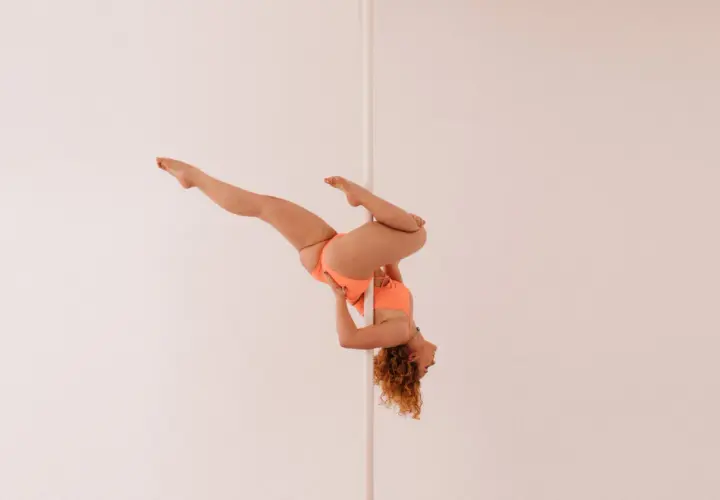 Kobieta w pomarańczowym stroju wykonuje skomplikowaną figurę na pionowym słupku, przypominającą flagę pole dance.