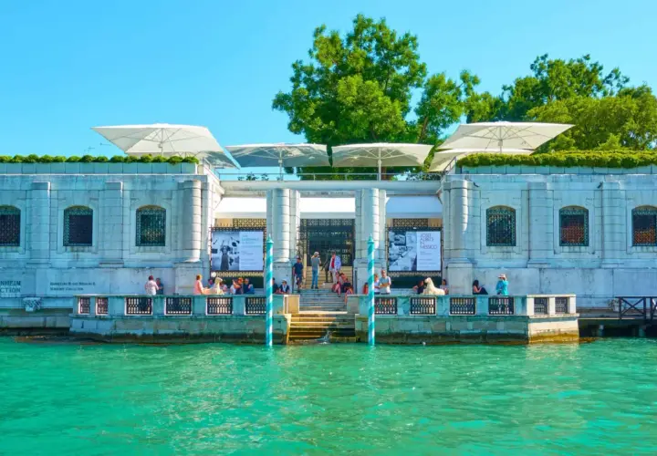 Le **Guggenheim Venise** se reflète dans l'eau turquoise. Des parasols blancs ombragent la terrasse où des visiteurs admirent l'architecture.