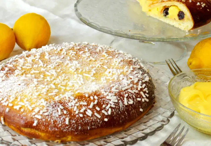 Una deliziosa polacca aversana, cosparsa di zucchero a velo e confettini, accanto a limoni freschi e una ciotola di crema pasticcera.