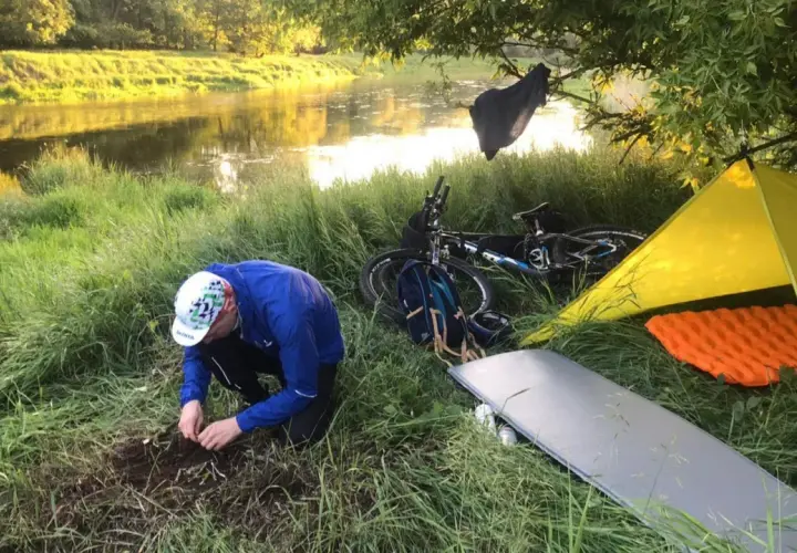 Biwak nad rzeką: rozłożony żółty namiot, materac, rower i mężczyzna przygotowujący ognisko. To idealny materiał na namiot i nocleg w plenerze.