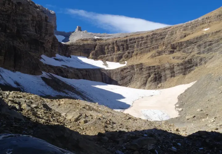 Un paisaje montañoso con nieve y rocas, donde el sol ilumina la **col de tentes** y el cielo azul.