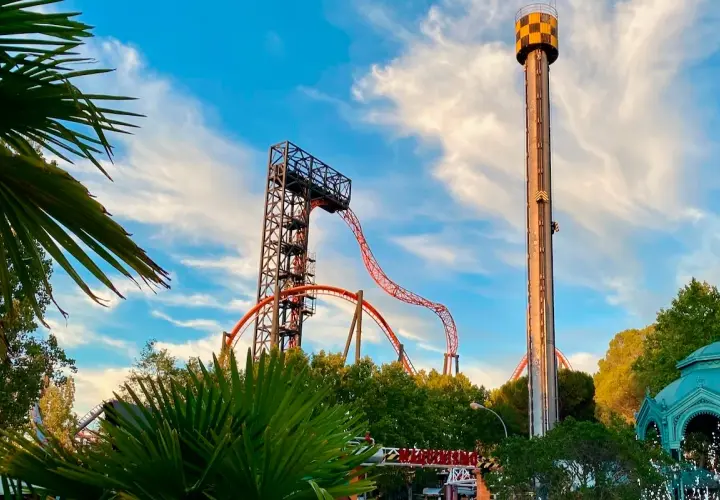 Torre de caída libre y montaña rusa anaranjada en un parque de atracciones con palmeras.