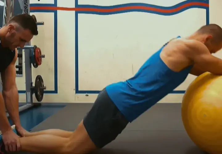 Trener pomaga wykonać ćwiczenie nordic curl, stabilizując nogi klienta opierającego się o dużą, żółtą piłkę gimnastyczną.