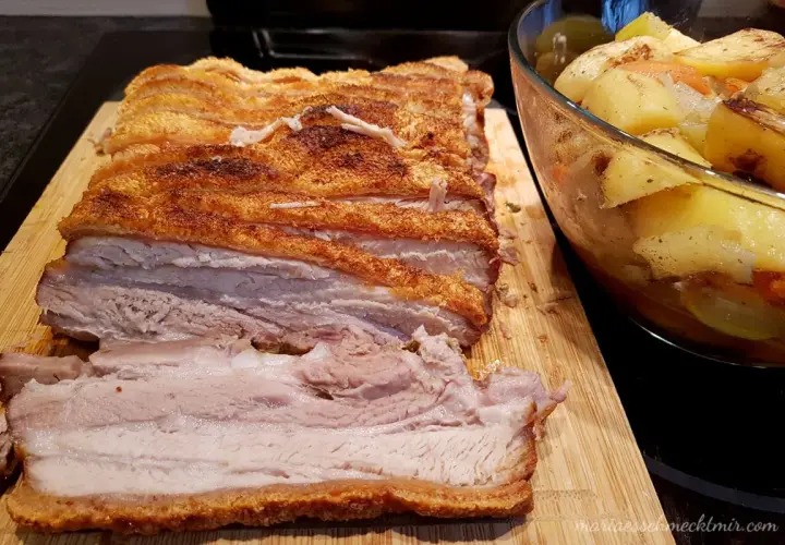 Ein butterzarter Schweinebauch mit knuspriger Kruste liegt auf einem Holzbrett, daneben eine Schüssel mit Bratkartoffeln und Gemüse.
