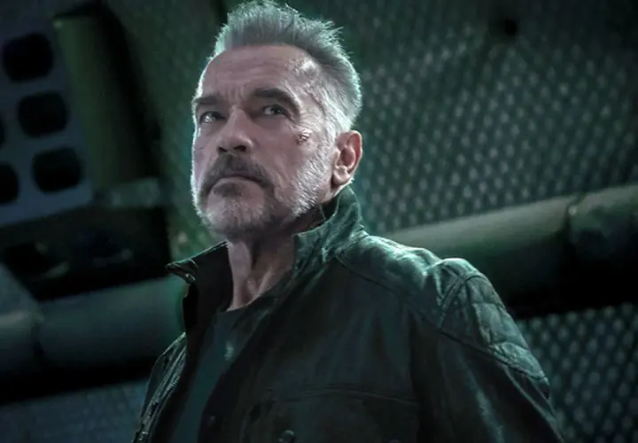 Arnold Schwarzenegger jako T-800 w filmach Jamesa Camerona. Z blizną na policzku, w skórzanej kurtce, patrzy w górę.