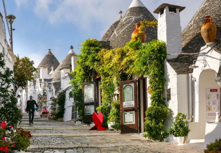 A person walks down a charming street lined with iconic trulli houses, lush greenery, and shops. This picturesque scene makes you wonder, which is the best town to stay in Puglia?