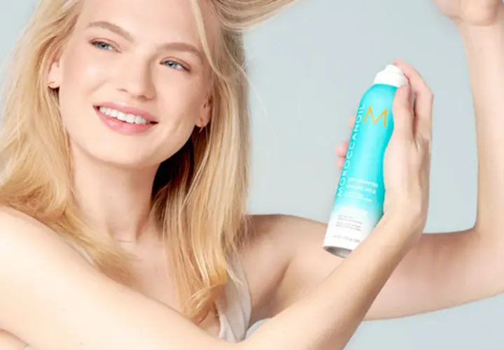 Mujer rubia sonriendo aplica champú seco para lavar pelo en seco, refrescando su melena.