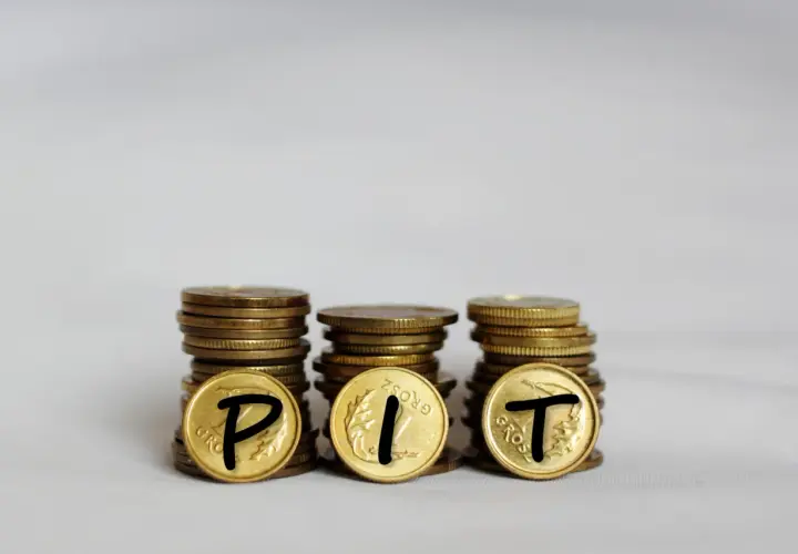 Trzy stosy monet, na których widnieją litery P, I, T. Symbolizują one rozliczenie podatku PIT.