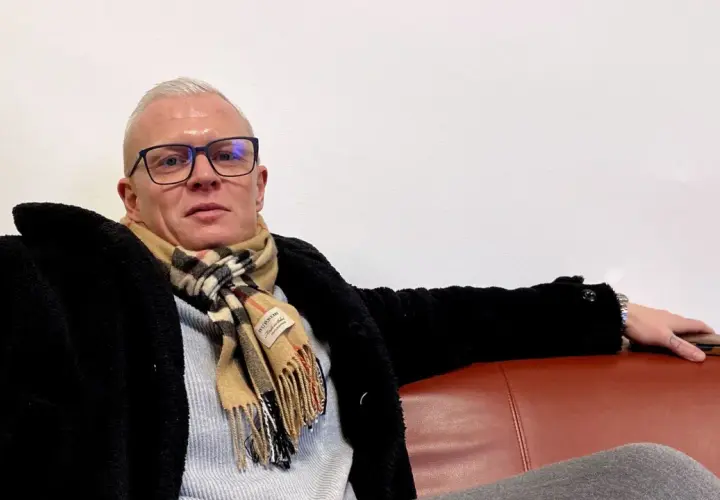 Robinson Alexander, med glasögon och en rutig halsduk, sitter avslappnat på en röd soffa.