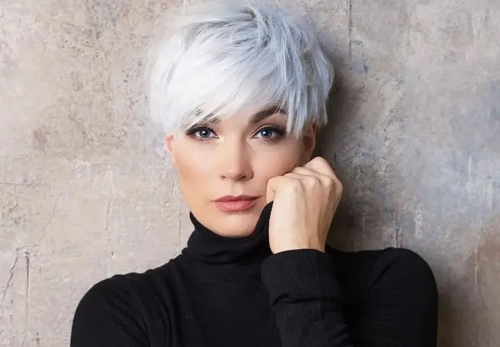 Kobieta z siwymi włosami w stylu pixie bob fryzura, ubrana w czarny golf, opiera dłoń o twarz.