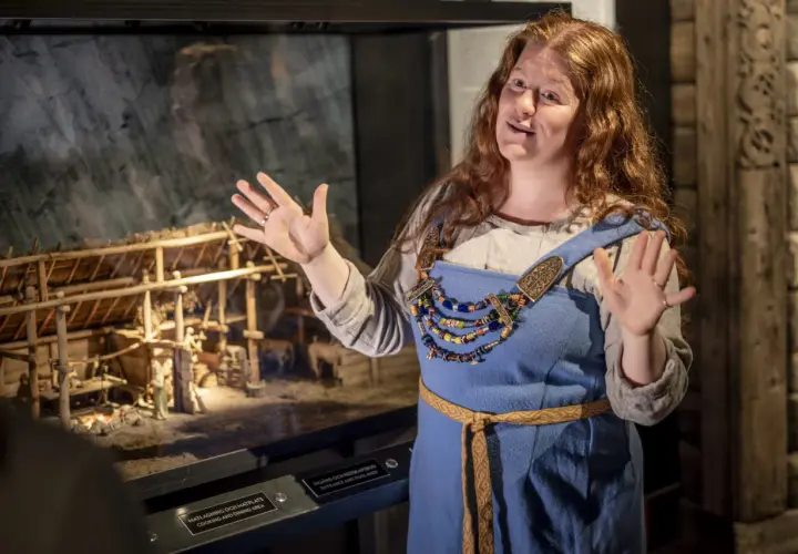 En kvinna i vikingakläder berättar vid en modell av en vikingagård på vikingamuseet.