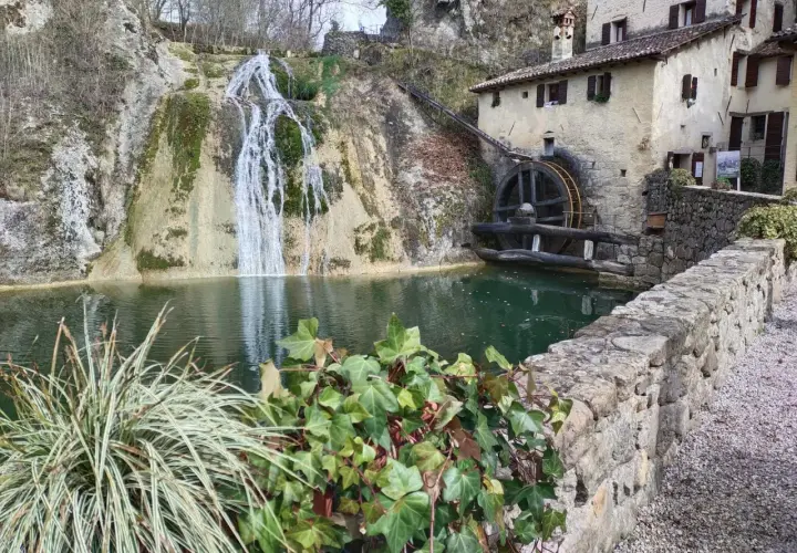 Antico mulino ad acqua accanto a cascate e grotte in Veneto, con un laghetto verde e vegetazione rigogliosa.